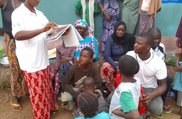 Causerie éducative de quartier : une animatrice présente un support illustré à des femmes, des jeunes et des enfants rassemblés devant une maison.