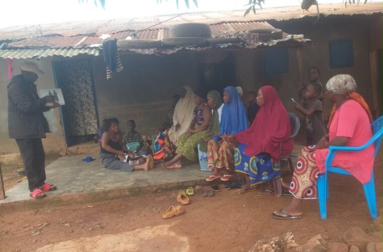 Causerie communautaire du projet PSR devant une concession, animée devant des femmes et des enfants assis sous l’auvent.