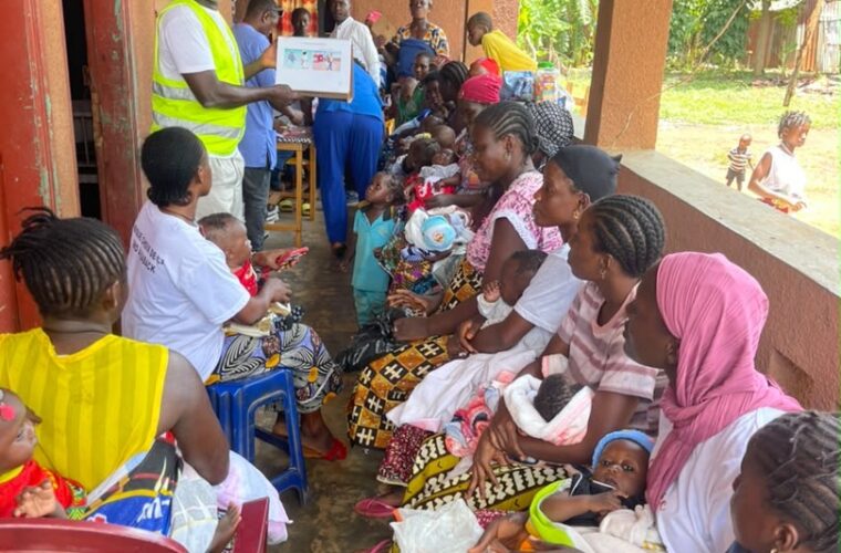 Séance de sensibilisation du projet PSR sous la véranda d’un poste de santé : un agent présente une boîte à images à des mères venues avec leurs enfants.