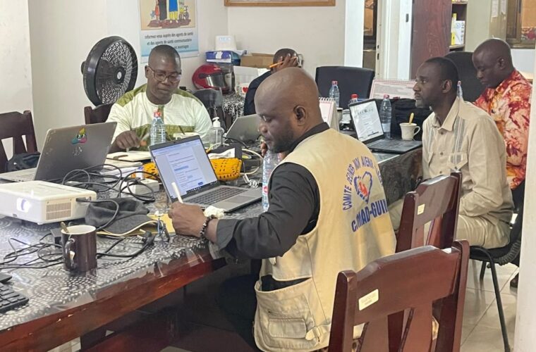 Séance de travail du projet RACINE : un membre de CJMAD Guinée, en gilet floqué au nom de l’organisation, prend des notes devant son ordinateur portable, entouré de collègues.